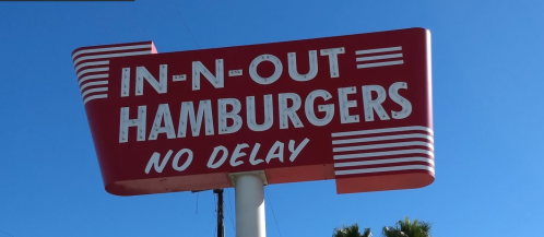 Sign for In-N-Out Burgers featuring the text "IN-N-OUT HAMBURGERS NO DELAY" against a clear blue sky.