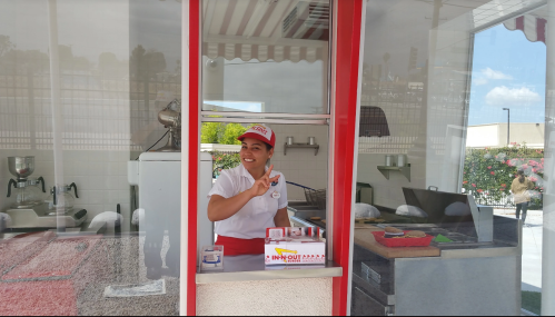 A smiling employee at a fast-food window, wearing a uniform and cap, giving a peace sign.