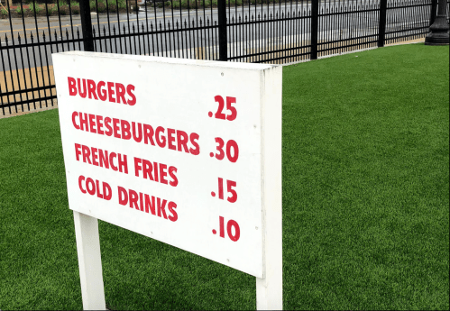 Sign displaying menu prices: Burgers $0.25, Cheeseburgers $0.30, French Fries $0.15, Cold Drinks $0.10 on green grass.