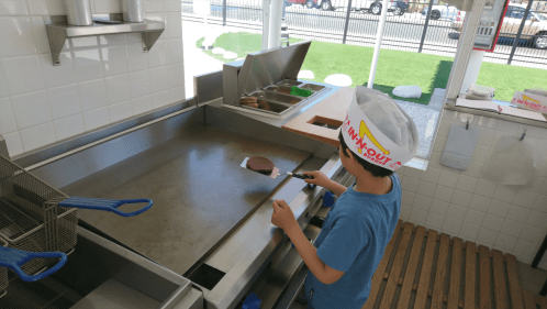 A child wearing a hat cooks a burger on a grill in a bright, clean kitchen.