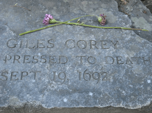 A stone grave marker inscribed with "GILES COREY PRESSED TO DEATH SEPT 19, 1692," adorned with a flower.