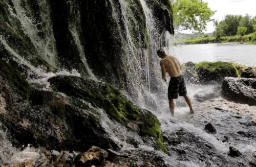 A person stands under a waterfall, surrounded by rocks and greenery, enjoying the cascading water.