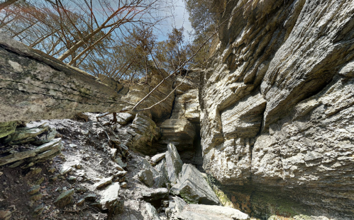 A rocky canyon with steep walls, scattered boulders, and sparse trees under a clear blue sky.