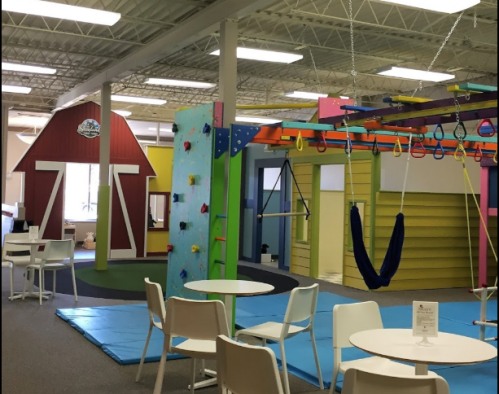 Bright indoor play area featuring climbing walls, swings, and colorful play structures, with tables and chairs for parents.