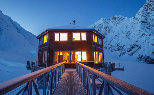 A cozy mountain cabin with glowing windows, surrounded by snow-covered peaks at dusk.