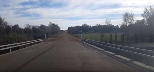 A dirt road leads over a bridge, surrounded by trees and fields under a partly cloudy sky.
