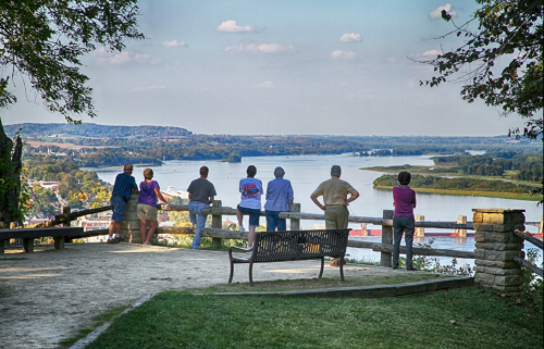 A group of people stands on a lookout, gazing at a river and scenic landscape under a clear sky.