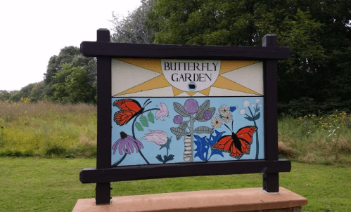 A colorful sign for a Butterfly Garden featuring butterflies and various flowers, set against a green landscape.