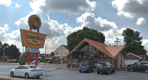 A diner with a distinctive sign, surrounded by cars and trees, under a partly cloudy sky.