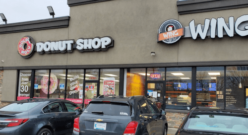 Exterior of a donut shop and wing restaurant, featuring bright signage and parked cars in front.