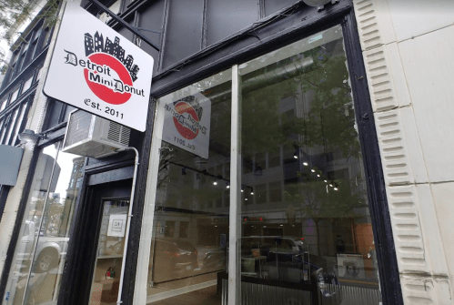 Exterior of Detroit Mini Donut shop, featuring a sign and large windows, showcasing a city vibe. Established in 2011.