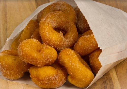 A paper bag overflowing with golden, sugar-coated donuts on a wooden surface.