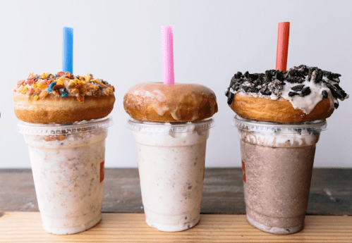 Three milkshakes topped with donuts: one with cereal, one glazed, and one with crushed cookies, each with a colorful straw.