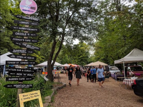A bustling outdoor market with vendors, tents, and visitors walking along a tree-lined path.