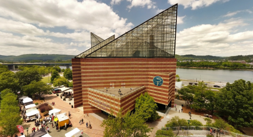 Aerial view of a modern building with a glass roof and striped brick facade, surrounded by greenery and a river.