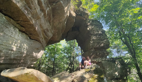 Two people sit on a large rock formation in a forest, surrounded by trees and sunlight filtering through the leaves.