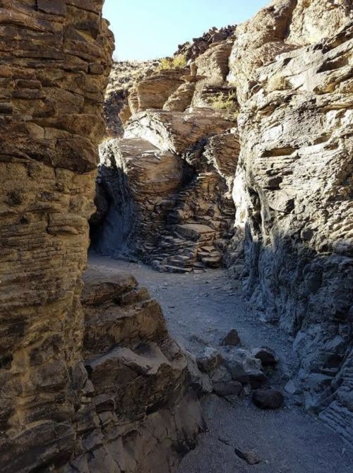 A narrow canyon with steep, rocky walls and a gravel path leading into the distance under a clear blue sky.