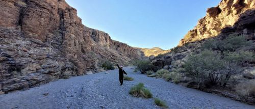 A person stands in a rocky canyon, surrounded by dry riverbed and sparse vegetation under a clear blue sky.