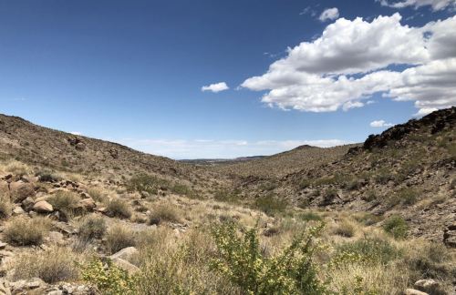 A wide view of a desert landscape with rocky hills, sparse vegetation, and a blue sky dotted with clouds.