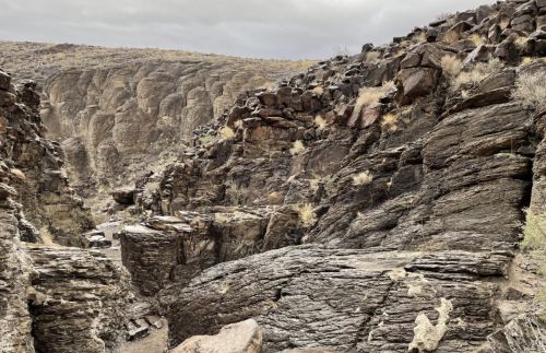 Rocky canyon landscape with layered stone formations and sparse vegetation under a cloudy sky.