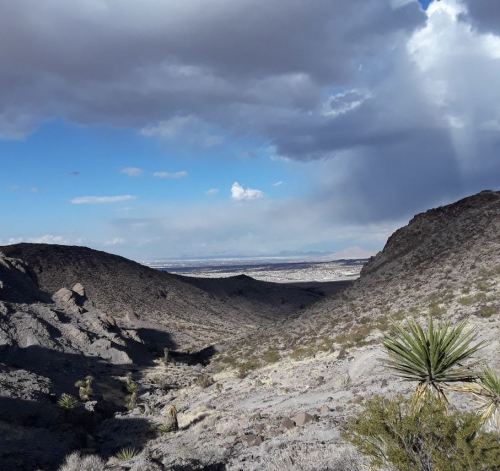 A scenic view of a rocky desert valley under a partly cloudy sky, with distant mountains and sparse vegetation.