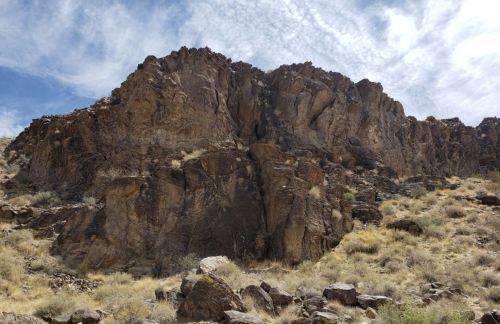 Rocky mountain landscape with rugged cliffs and sparse vegetation under a cloudy blue sky.