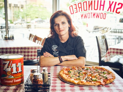 A chef sits at a table with a large pizza and a can of tomatoes, showcasing a cozy restaurant setting.