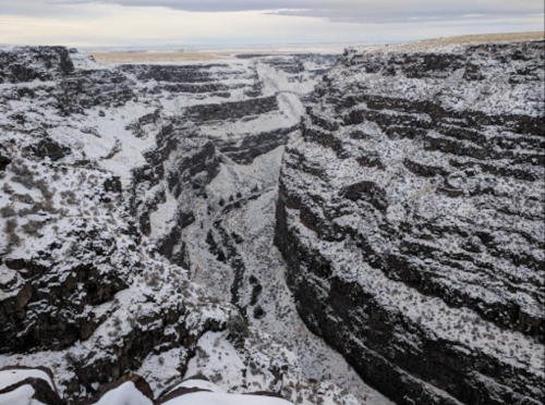 A snowy canyon landscape with steep, rocky walls and a winding river at the bottom, under a cloudy sky.