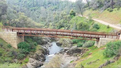 A wooden bridge spans a rocky creek, surrounded by lush greenery and trees, with a dirt path in the background.