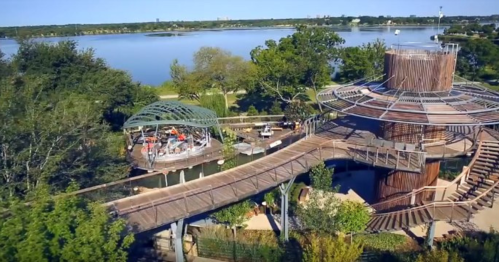 Aerial view of a modern wooden structure with a walkway, surrounded by trees and a lake in the background.