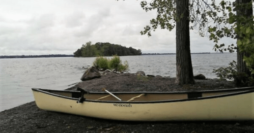 A canoe on a rocky shore by a calm lake, surrounded by trees and a cloudy sky.