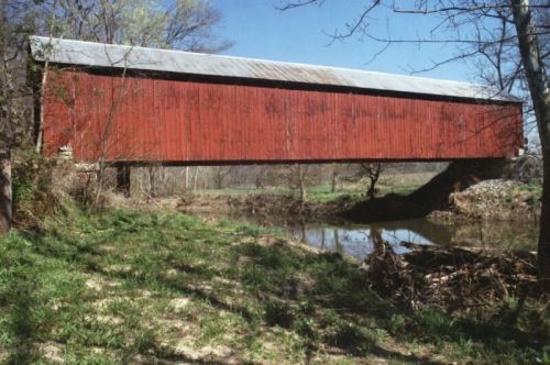 A red covered bridge spans a small stream, surrounded by green grass and trees under a clear blue sky.