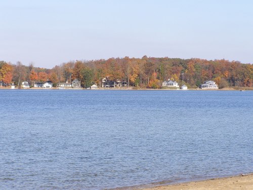 A serene lake scene with colorful autumn trees and houses along the shore under a clear blue sky.