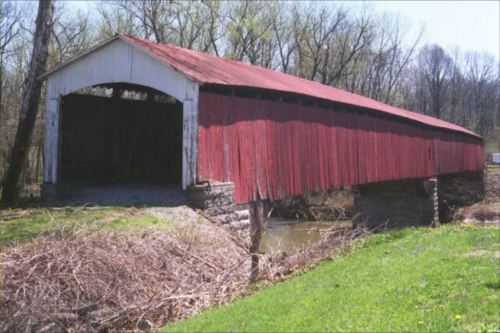 A red covered bridge with a white roof spans a small stream, surrounded by greenery and trees in the background.