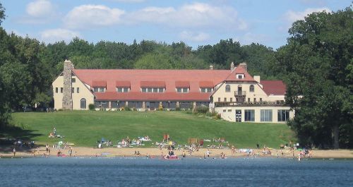 A large building with a red roof overlooks a sandy beach filled with people enjoying a sunny day by the water.