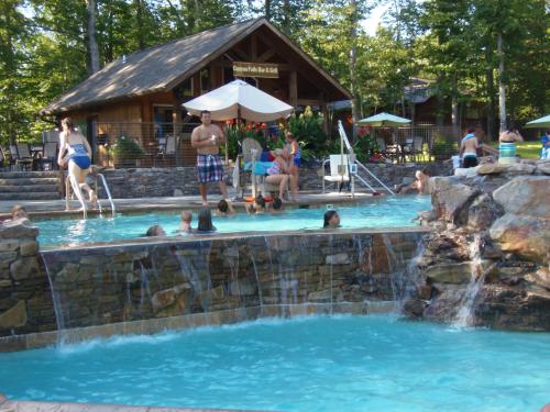 A sunny pool scene with people swimming and relaxing, surrounded by trees and a rustic lodge in the background.