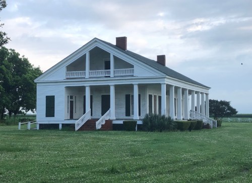 A large white house with a porch, surrounded by green grass and trees, under a cloudy sky.