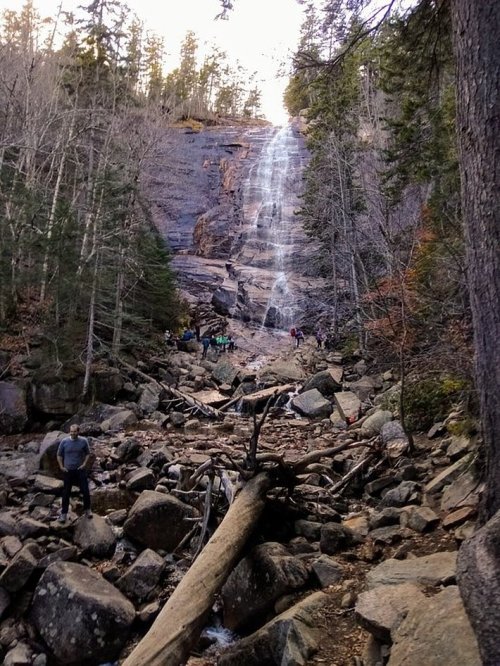 A waterfall cascades down rocky cliffs, surrounded by trees and hikers exploring the rocky terrain below.
