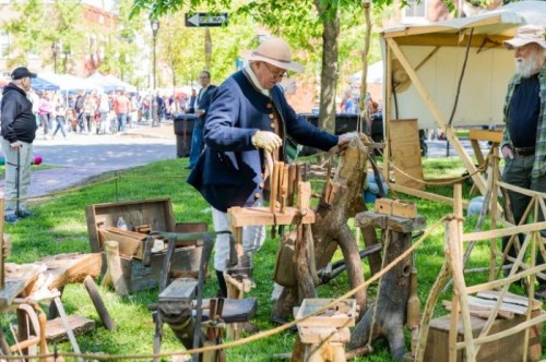 A craftsman in period attire demonstrates woodworking at an outdoor festival, surrounded by handmade wooden tools and furniture.