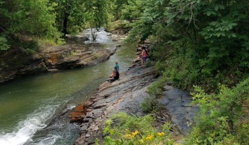 Two people sit on rocky riverbank surrounded by lush greenery, with a gentle stream flowing nearby.