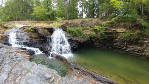 A serene waterfall cascades over rocky terrain into a calm green pool, surrounded by lush trees and foliage.