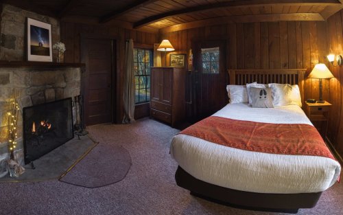 Cozy bedroom with a wooden interior, a fireplace, and a bed adorned with a red and white blanket. Warm lighting creates a welcoming atmosphere.