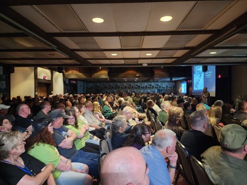 A large audience sits in a conference room, attentively listening to a speaker on stage.