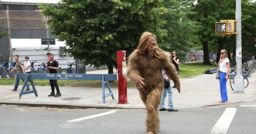 A large, furry creature resembling Bigfoot walks through a city street, with people observing nearby.