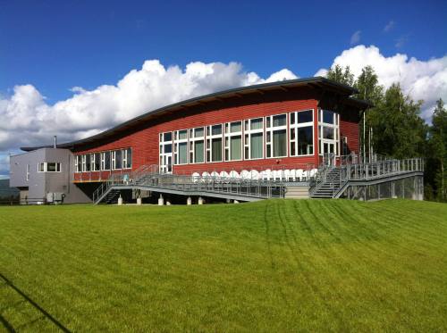 Modern building with large windows, a curved roof, and a deck, set on a green lawn under a blue sky with clouds.