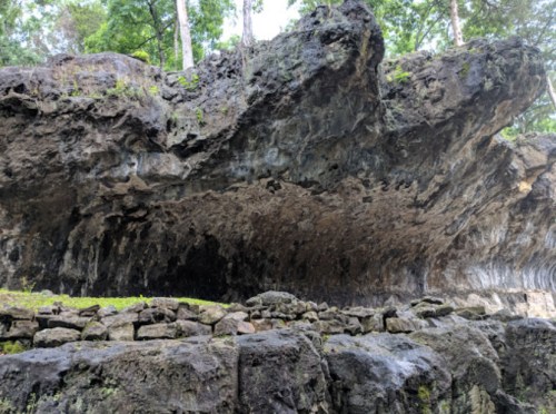 A rocky cliff with overhanging formations, surrounded by lush greenery and a stone wall at the base.