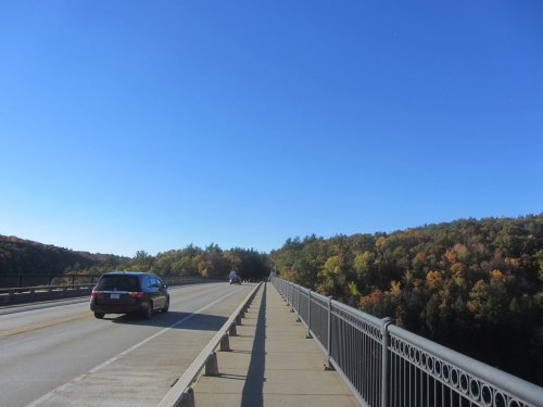 A car drives on a bridge with a clear blue sky and colorful trees in the background.