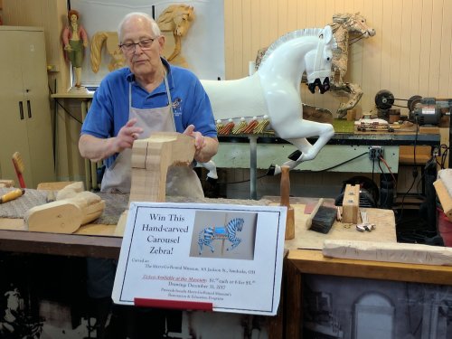 An elderly man carves wood at a workshop, with a sign promoting a hand-carved carousel zebra in the background.