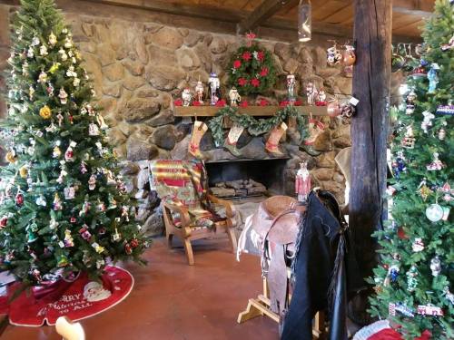 Cozy holiday scene with decorated Christmas trees, a stone fireplace, and festive decorations in a rustic setting.