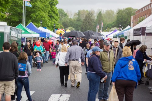 A busy street fair with people walking among colorful tents and umbrellas on a cloudy day.